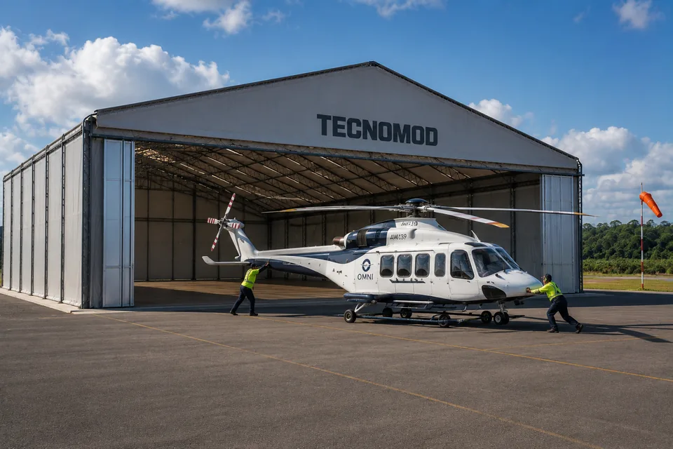 Galpão lonado em uso como hangar de helicóptero em aeroclube, com a testeira inteira aberta e a aeronave sendo recolhida