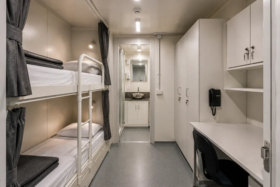 Interior de módulo offshore de acomodação, com beliches, cortinas de privacidade, armários individuais, escrivaninha e banheiro ao fundo